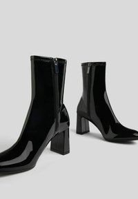 Czarne lakierowane botki za kostkę na kwadratowym obcasie, z boku zapinane na zamek błyskawiczny, o gładkiej, błyszczącej strukturze. Zaprojektowane w smukłej sylwetce.