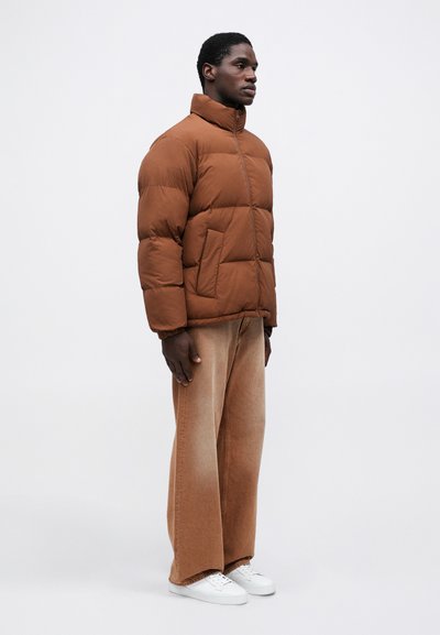 Brun polstret jakke med høj krave, der har en frontlynlås og to sidelommer; båret med lysebrune brede jeans og hvide sneakers.