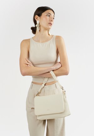 Donna che indossa un top beige senza maniche e pantaloni, grandi orecchini bianchi, tiene una borsa bianca con manici intrecciati attraversata sul corpo.