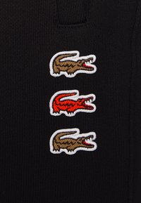 Svart tyg med tre broderade krokodil-logotyper i brunt och rött, omramade i vitt. Texturen är ribbad och ger en unik detalj.