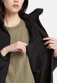 Giacca nera con zip frontale, polsini elasticizzati e una tasca. T-shirt color verde oliva sotto, con una texture sottile e un piccolo logo.