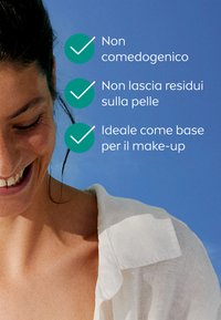 Maglietta bianca su una persona con i capelli scuri, sorridente. Testo su sfondo blu elenca i benefici del prodotto: non comedogenico, senza residui, base ideale per il trucco.