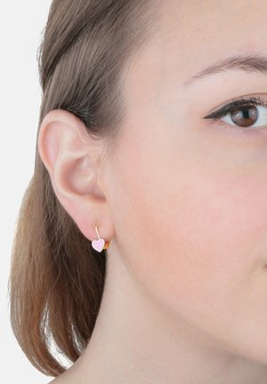 Ohr mit kurzen braunen Haaren, das einen goldenen Ohrring mit einem kleinen rosa Herzanhänger trägt, vor einem schlichten weißen Hintergrund.
