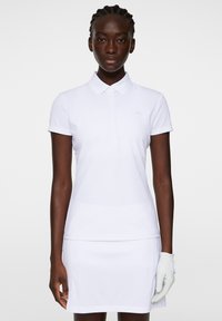 Vit pikétröja med krage och korta ärmar, med en ljusrosa logotyp på bröstet. Slät textur, kombinerad med en vit kjol.