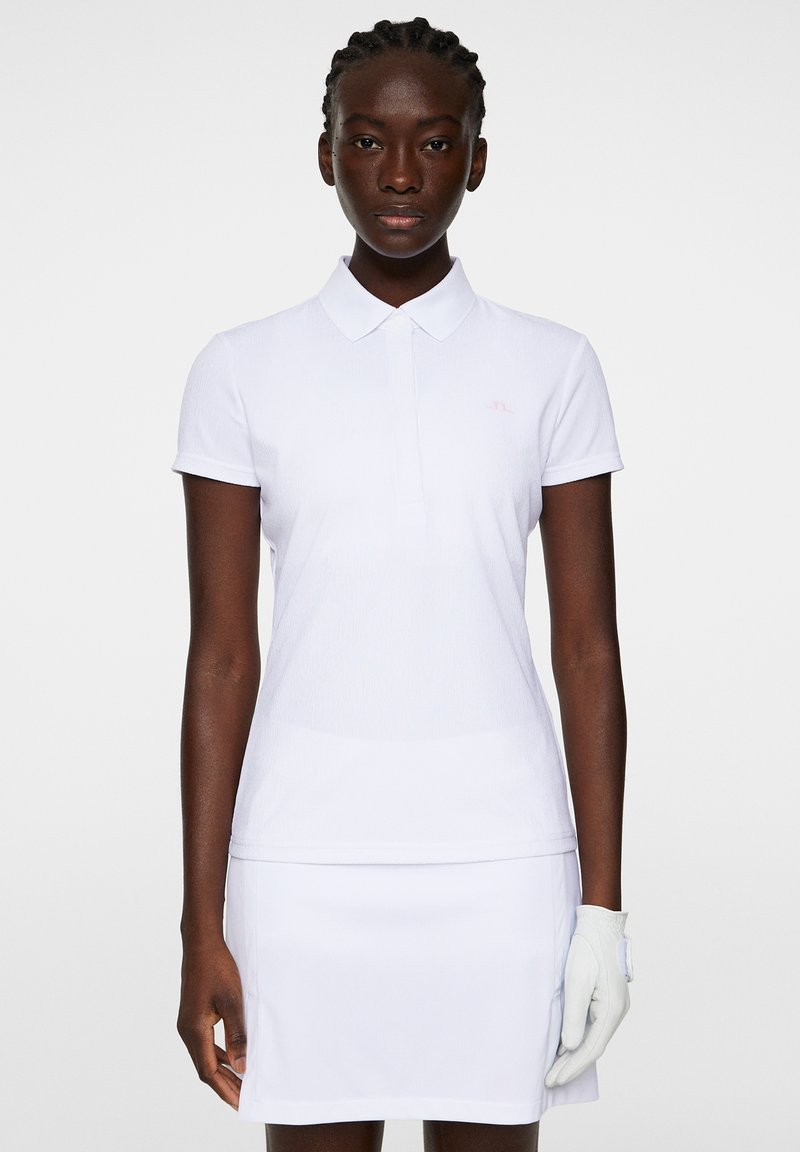 Vit pikétröja med krage och korta ärmar, med en ljusrosa logotyp på bröstet. Slät textur, kombinerad med en vit kjol.