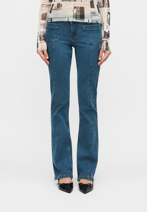 Jean bleu à jambes droites avec des poches avant plaquées, porté avec des chaussures noires à bout pointu et un haut à manches longues imprimé de motifs de journaux.