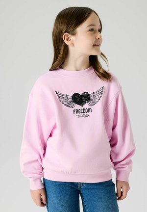 Fille portant un sweat-shirt rose avec un cœur ailé noir et le texte « FREEDOM World Tour », associée à un jean bleu, regardant vers la droite et souriant.