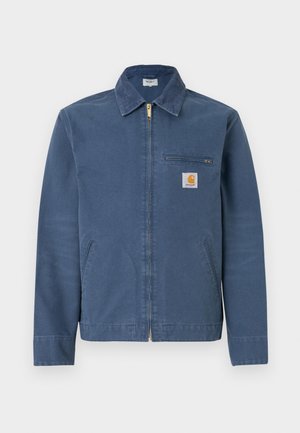 Blå lærred zip-up jakke med krave, lommer foran og en brystlomme med et mærkets logo patch. Guld lynlåsdetalje.