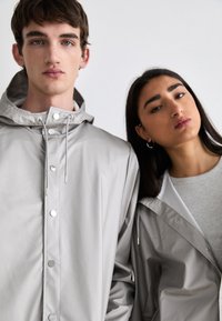Vestes imperméables gris clair avec capuche, présentant des fermetures à pression, des cordons de serrage ajustables et une texture lisse, mate. Coupe moyenne pour un usage décontracté.