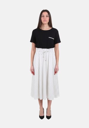 Donna con capelli lunghi e lisci in piedi di fronte, indossa una maglietta nera a maniche corte e una gonna bianca a vita alta con sottili righe verticali e tacchi neri.