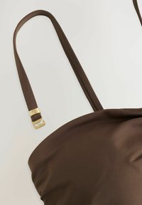 Top in nylon marrone con spalline sottili e una fibbia in metallo dorato. Texture liscia con un design leggermente arricciato alla base.