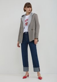 Blazer à carreaux avec un sweat à capuche gris, un t-shirt blanc, un jean foncé taille haute et des mules à talons rouges. Les accessoires incluent de petites boucles d'oreilles.