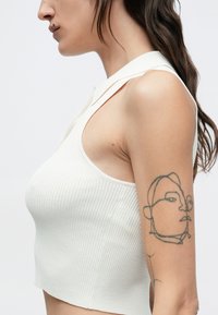 Biely rebrovaný krátky top s hrotovým golierom, ktorý má otvorený dizajn na ramene. Na hornej časti paže je viditeľné tetovanie v štýle kresby.