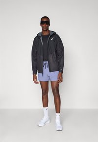 Leichte schwarze Herrenjacke mit Kapuze, kombiniert mit einem schwarzen T-Shirt und helllila Shorts. Accessoires sind Sonnenbrillen und weiße Sneaker.