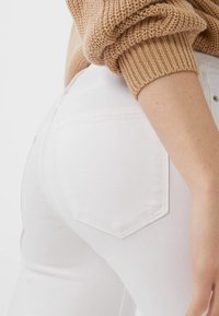 Manga de suéter beige de punto sobre el bolsillo del vaquero blanco en la parte baja de la espalda y el brazo de una persona, con un fondo liso.