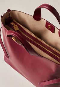 Burgundy leather tote bag with a smooth texture, featuring a zippered interior pocket and a light-colored suede lining. Gold-tone hardware accents.