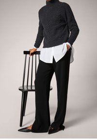 Personne portant un pantalon large noir, des chaussures pointues noires, une chemise blanche et un pull en maille torsadée gris foncé, debout à côté d'une chaise en bois noire.