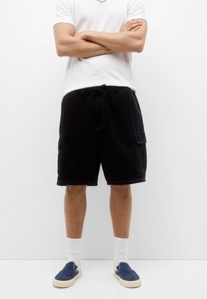 Black cargo shorts with a drawstring waist, featuring side pockets and a relaxed fit. Paired with a white t-shirt and navy slip-on shoes.