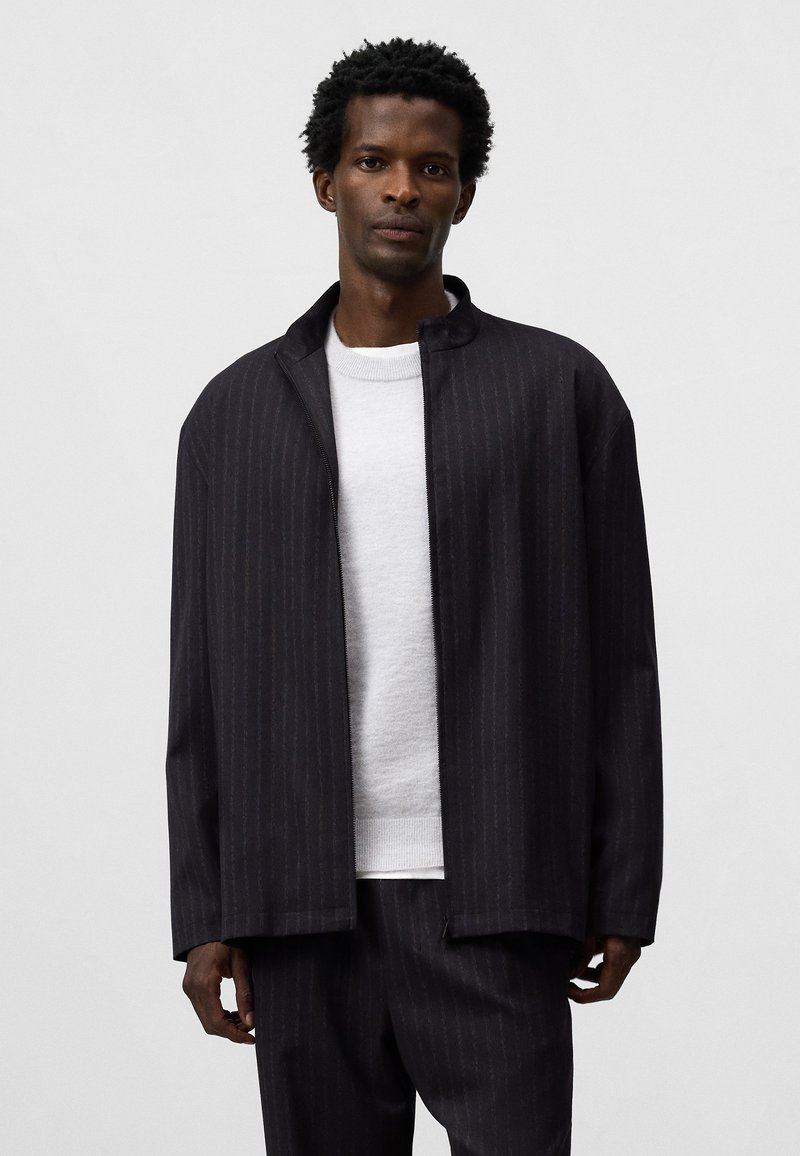 Giacca nera a righe sottili con una vestibilità rilassata, dotata di colletto alto e texture liscia, indossata sopra un maglione grigio chiaro.