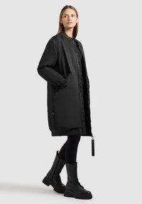 Zwarte gewatteerde lange jas met een opstaande kraag, ritssluiting, twee zijzakken en geribbelde manchetten. Draag over zwarte leggings en zwarte veterschoenen.
