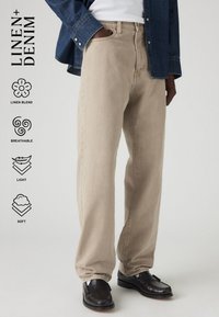 Beige vászon keverék anyagú farmernadrág egyenes szabású, sima textúrával és oldalsó zsebekkel, sötét mokaszin cipőkkel és farmerekinggel párosítva.