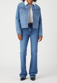 Chaqueta vaquera con cuello de piel, color azul, dos bolsillos en el pecho y detalles de botones, combinada con pantalones vaqueros acampanados azules y botas negras.