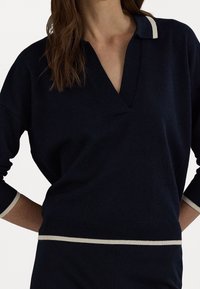 Nainen, jolla on yllään tummansininen pitkähihaisen neulepaita, jossa on V-kaula-aukko, kaulus ja beige reunus kauluksessa, hihoissa ja helmassa, valkoista taustaa vasten.