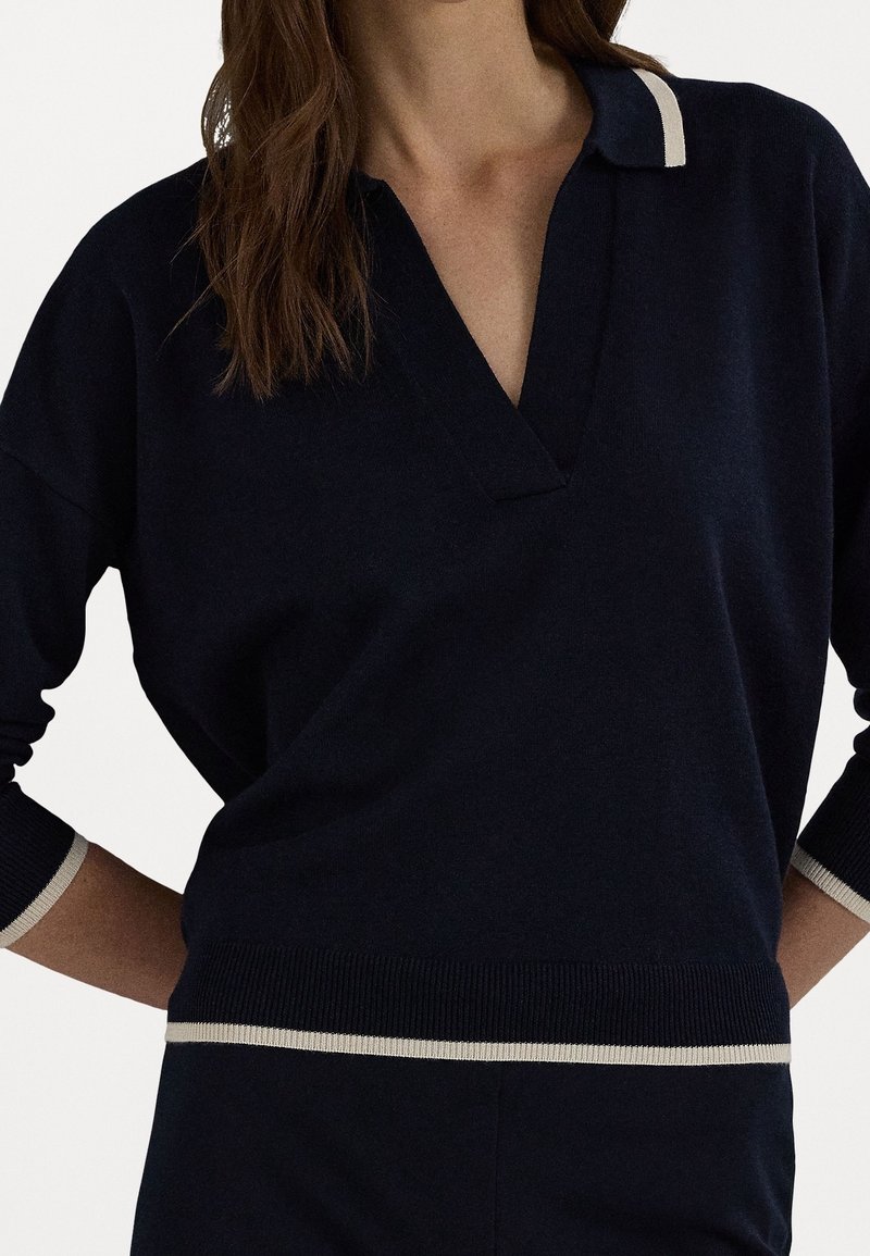 Nainen, jolla on yllään tummansininen pitkähihaisen neulepaita, jossa on V-kaula-aukko, kaulus ja beige reunus kauluksessa, hihoissa ja helmassa, valkoista taustaa vasten.