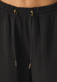 Black drawstring pants made from textured fabric. Features elastic waistband with gold-toned hardware accents on drawstrings.