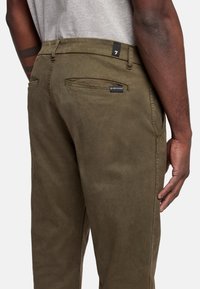 Pantaloni verde oliva con una consistenza liscia, dotati di tasche posteriori e un sottile etichetta del marchio vicino alla vita. Design con fronte piatto e taglio dritto.