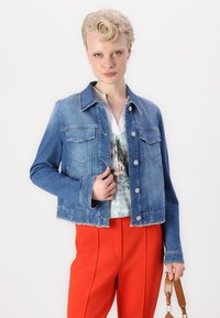 Jeansjacke in Mittelblau mit cropped Design, Knopfverschluss, Brusttaschen und rohem Saum, kombiniert mit einem gemusterten Oberteil und roten Hosen.