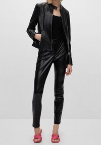 Kobieta w czarnej skórzanej kurtce i spodniach, z czarnym topem, stoi z rękami w kieszeniach, w różowych sandałach na wysokim obcasie z odkrytymi palcami na jednolitym tle.