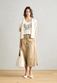 Beige A-linje skjørt med frontknapper, hvit t-skjorte med et fargerikt grafisk motiv, og en strikket kremfarget cardigan. Kombinert med hvite pumpesko og tote-veske.