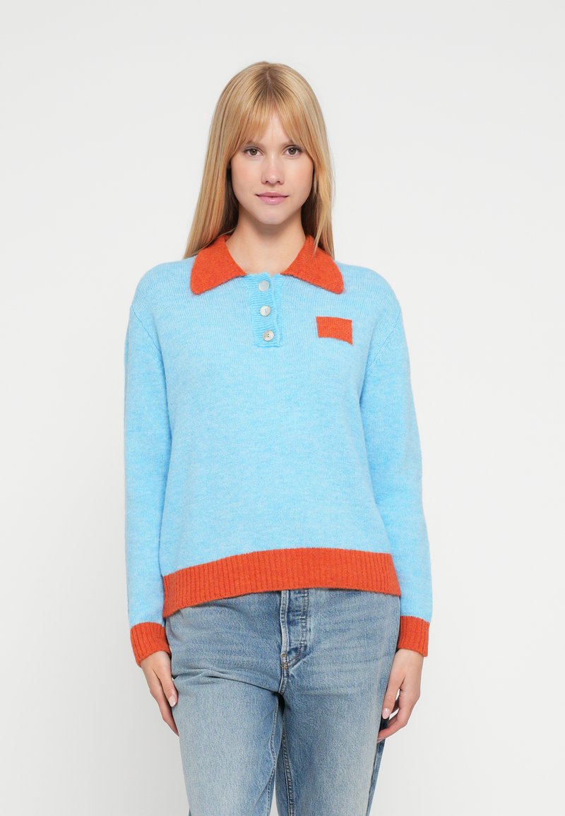 Hellblauer Pullover mit einem orangefarbenen Rippenkragen und Saum, versehen mit drei Knöpfen und einer kleinen Brusttasche in demselben Orange.
