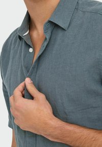 Camisa de manga corta abotonada en tejido de lino color gris pizarra. Presenta un cuello puntiagudo, cierre de un solo botón y una textura sutil.