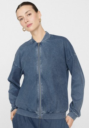 Frau mit goldenen Creolen, die eine verblasste blaue Bomberjacke mit Reißverschluss und passende blaue Hosen trägt, vor einem schlichten Hintergrund stehend.