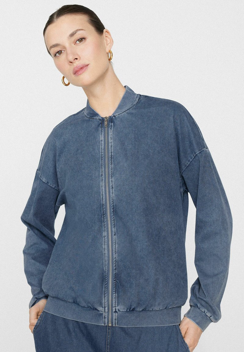 Kobieta z złotymi kolczykami kołami, ubrana w wyblakłą, niebieską kurtkę bombkę na zamek błyskawiczny i pasujące niebieskie spodnie, stojąca na jednolitym tle.