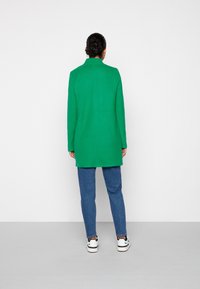 Abrigo verde de corte recto y cuello alto, hecho de un tejido texturizado, combinado con vaqueros azules y zapatillas blancas.