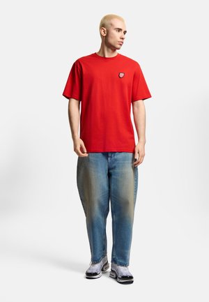 Młody mężczyzna z krótkimi blond włosami, ubrany w czerwony t-shirt z krótkim rękawem, luźne wyblakłe niebieskie dżinsy i białe trampki, stoi na jednolitym tle.