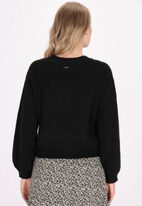 Schwarzer Strickpullover mit einem strukturierten Muster, gerippten Bündchen und Saum; verfügt über ein kleines Metall-Logo am hinteren Halsausschnitt.