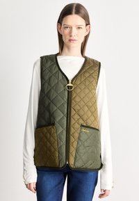 Πράσινο γιλέκο καπιτονέ με δύο μπροστινές τσέπες, χρυσό φερμουάρ και αντίθετη ελιά πινακίδα. Λογότυπο στην τσέπη και εμφανές υφασμάτινο μοτίβο.