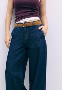 Kä widerleggende denimbukser i mørkeblå, med et brunt lærbelte. Stoffet har en glatt tekstur med minimal sømdetalj.