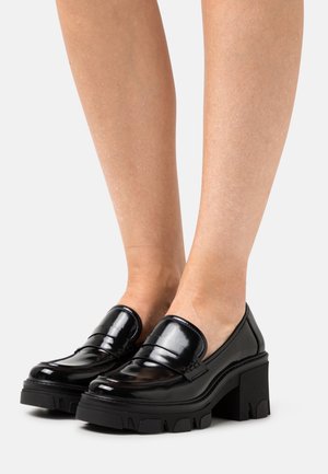 Mocasines de charol negro brillante con tacones robustos, usados sobre piernas desnudas, con suelas gruesas y redondeadas.