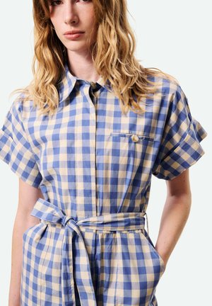 Femme portant une robe ceinturée à carreaux bleus et beiges, à manches courtes, avec une poche poitrine boutonnée et des poches latérales, debout avec une main dans la poche.