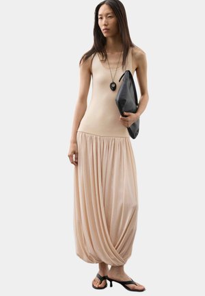 Kvinde med langt hår iført en ærmeløs beige kjole, sorte hælsandaler, en sort vedhængshalskæde og holder en stor sort clutch.