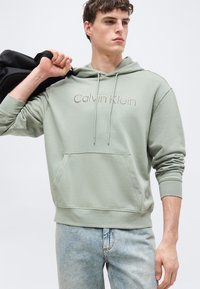 Jeune homme portant un sweat à capuche Calvin Klein vert clair et un jean, tenant une veste noire sur son épaule, regardant sur le côté.