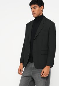 Blazer preto com um único botão, lapelas com corte em bico e bolso no peito. Usado por cima de um pulôver de gola alta preto, combinado com jeans cinzentos.