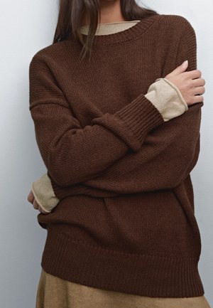 Kvinde med armene krydset iført en lagdelt mørkebrun strikket sweater over en beige langærmet skjorte mod en ensfarvet lys baggrund.
