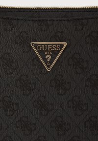 Sac en tissu noir structuré arborant un subtil motif embossé. Plaque logo triangulaire dorée avec "GUESS USA" affichée de manière proéminente.