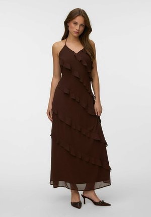 Mujer con un vestido largo marrón sin mangas con capas de volantes diagonales y tacones marrones puntiagudos sobre un fondo liso claro.
